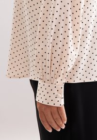 Lange mouwblouse in crème met zwarte stippen, voorzien van eenGathered cuffs met een knoopdetail. Zachte, gladde stoftextuur.