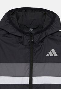 Jachet�ă neagră cu glugă, cu fermoar frontal, având dungi orizontale gri și alb și un logo reflectorizant Adidas pe piept.