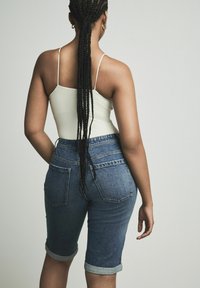 Pantalones cortos de mezclilla azul oscuro con dobladillos enrollados, diseño de cintura alta y bolsillos traseros; combinados con una camiseta de tirantes blanca ajustada.