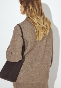 Brūns adīts džemperis ar augstu apkakli un ribotām malām, apvienots ar tumši brūnu zamša rokassomu, redzams no aizmugures. Redzama mīksta faktūra.