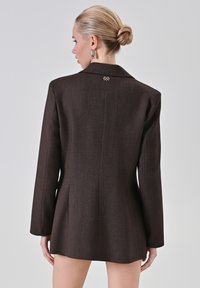 Blazer marrone su misura in tessuto a spina di pesce testurizzato, con maniche lunghe e un accento dorato con logo sul colletto posteriore.