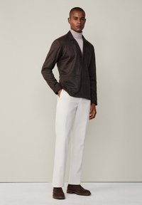 Giacca strutturata marrone scuro con due tasche frontali, pantaloni bianchi a sigaretta e scarpe di pelle marroni. Modello che indossa un dolcevita chiaro.