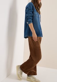 Donkerblauwe denim shirt, knopen dicht, met opgerolde mouwen; bruine corduroy wijde broek; beige sneakers met lichte accenten.