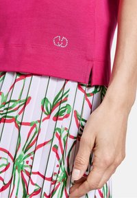 Camiseta rosa con un logo decorativo de strass en el dobladillo, combinada con una falda blanca plizada que presenta patrones florales en rojo y verde.