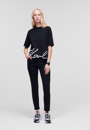 Schwarzes Baumwoll-T-Shirt mit weißem "Karl"-Logo am Saum, kombiniert mit schwarzen gemusterten Hosen und schwarz-weißen Sportschuhen.