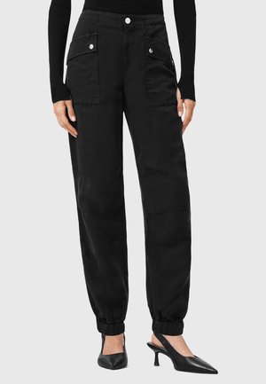 Zwarte cargo-stijl broek met elastische boorden, voorzakken, knoopsluiting en een gladde textuur. Gecombineerd met zwarte puntige hakken.
