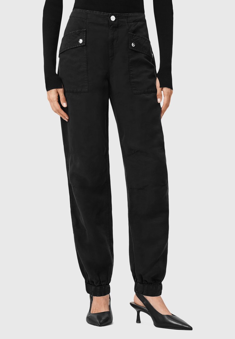Zwarte cargo-stijl broek met elastische boorden, voorzakken, knoopsluiting en een gladde textuur. Gecombineerd met zwarte puntige hakken.