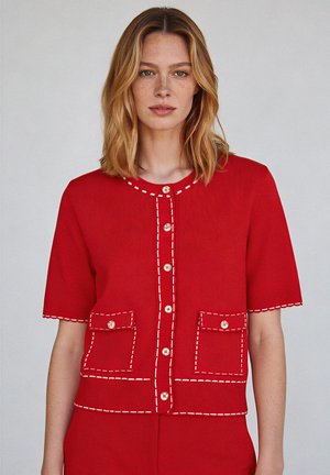 Vrouw met schouderlang blond haar die een rood gebreid vest met korte mouwen draagt, voorzien van witte stiksels en borstzakken.