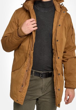 Man draagt een bruine parka-jas over een donkergrijze coltrui en olijfgroene broek, en past de rits van de jas aan met zijn linkerhand.