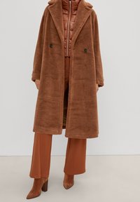 Abrigo largo de teddy marrón sobre una chaqueta acolchada marrón con cremallera, pantalones de pierna ancha y botas de ante de punta en una persona frente a un fondo liso.