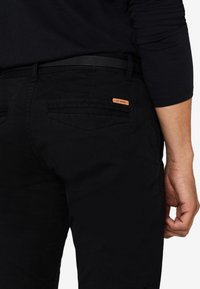 Jean noir en denim avec une coupe ajustée, une texture subtile, deux poches à l'arrière et un petit patch en cuir brun sur le côté droit.