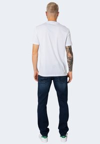 Tricou alb cu maneci scurte, blugi bleu și adidași albi cu accente verzi. Tatuaj vizibil pe brațul stâng. Fundal simplu.