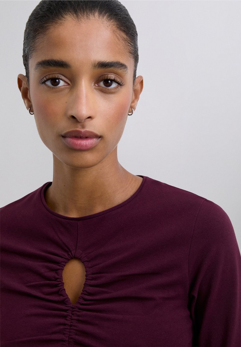 Burgundy fitted top with a round neckline, featuring a central keyhole cutout and gathered detailing beneath. Smooth texture, long sleeves.