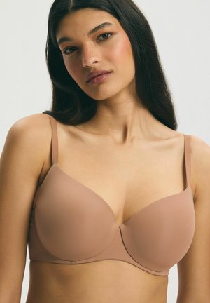 Woman with long dark hair wearing a smooth, light brown bra with thin straps against a plain light background.