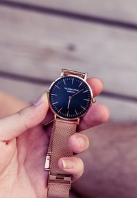Rose gold mesh strap wristwatch with a black face, slim hour markers, and a minimalistic design. Brand name visible on the dial.