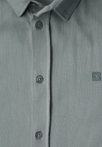 Ljusgrå skjorta med krage, prydd med fem mörkgrå knappar och ett litet logopatch på tyget.