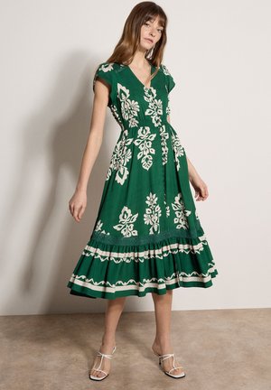 Kobieta pozuje w zielonej sukience midi z białymi kwiatowymi wzorami i guzikami, nosząc białe sandały na obcasie z paskami.