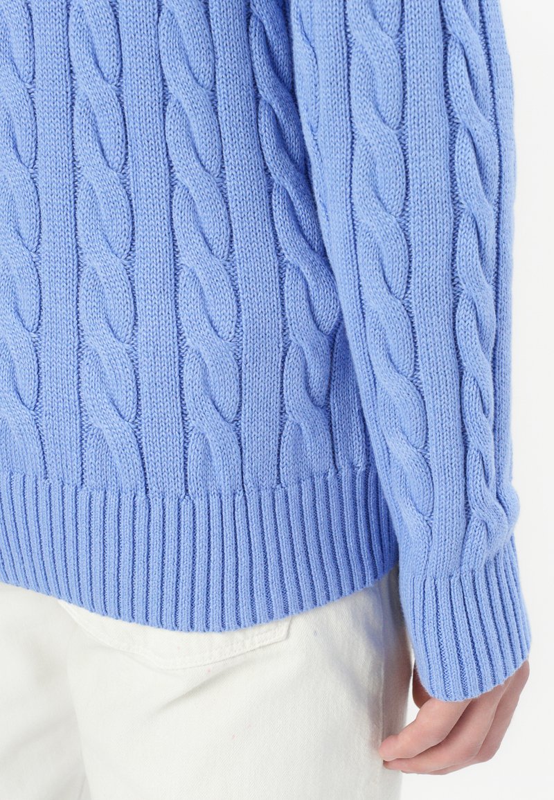 Maglione a maglia cable in azzurro chiaro, con motivi testurizzati e polsini e orlo a coste. Indossato sopra pantaloni bianchi, con dettagli delle mani in vista.