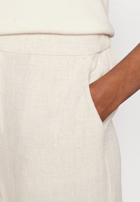 Pantalones de lino beige con un corte relajado, que cuentan con una cinturilla elástica y bolsillos laterales. La tela tiene una apariencia texturizada y tejida.