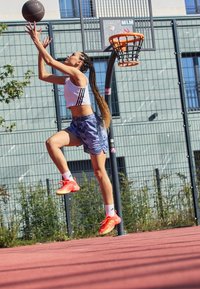 Basketbalista v bílém zkráceném topu a modrých šortkách se vzorem skáče, aby vystřelil míč, na nohou má jasně oranžové boty a bílé ponožky.