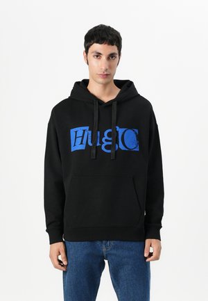 Svart hettegenser med frontlomme, snor i hetten, og en fet blå "Hugo" logo. Stoffet ser glatt ut med en avslappet passform. Bært med blå jeans.