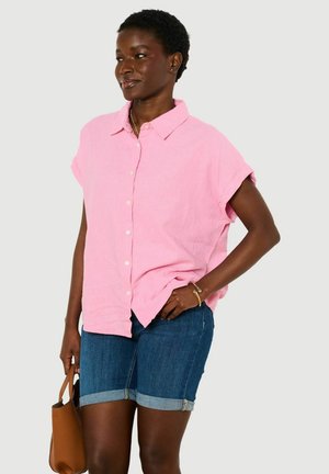 Kobieta stojąca, ubrana w luźną różową koszulę na krótki rękaw z guzikami, niebieskie podwinięte krótkie dżinsy, trzymająca brązową torebkę.