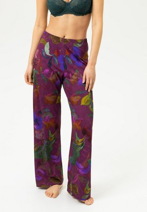 Femme portant un pantalon violet taille haute à jambes larges avec un imprimé floral coloré, associé à un soutien-gorge en dentelle vert foncé, pieds nus sur fond blanc.