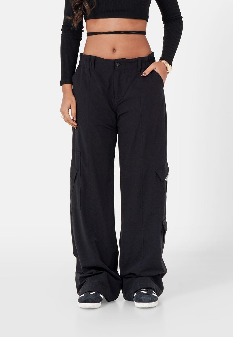 Zwarte wijde cargo broeken met diepe zijzakken, knoopsluiting en elastische tailleband. Gedragen met een cropped zwart top en zwarte sneakers.