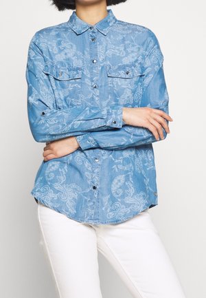 Person wearing a blue paisley-patterned button-up shirt with long sleeves crossed, paired with white pants on a plain background.