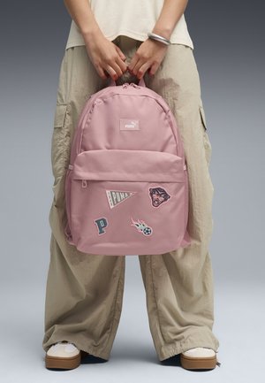 Person, der holder en lyserød Puma-rygsæk med patches, iført beige bukser med vide ben og hvide sneakers mod en grå baggrund.