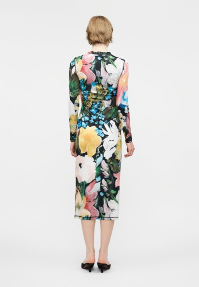 Άτομο με κοντά ξανθά μαλλιά που φοράει μακρυμάνικο, μεσαίου μήκους φόρεμα με μεγάλα πολύχρωμα λουλούδια, στέκεται με την πλάτη στην κάμερα και μαύρες γόβες.