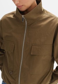 Olivgrüne Bomberjacke aus leichtem Material. Verfügt über einen hohen Kragen, einen Frontreißverschluss und zwei Brusttaschen für zusätzlichen Nutzen.