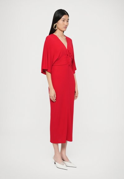 Sarkanā apvijanā kleita ar īsām, platiem piedurknēm, savilktu vidukļa daļu ar loku un grīdas garuma svārkiem. Kombinēta ar baltām kurpēm ar norādītām purngalām.