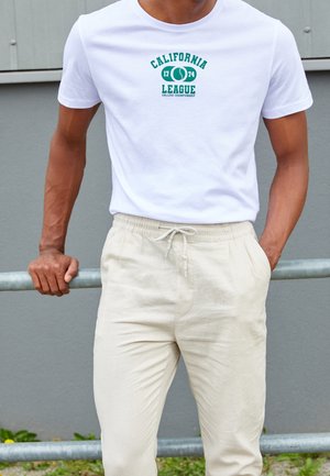 Homme portant un T-shirt blanc "California League 1774 College Championship" et un pantalon beige à cordon, appuyé contre une rambarde en métal en plein air.