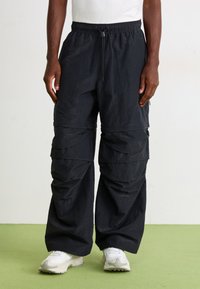 Mand i sorte, brede cargobukser med flere lommer og hvide sneakers, stående på et grønt gulv mod en enkel væg.
