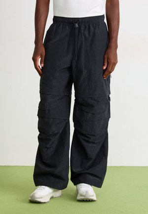 Man wearing black wide-leg cargo pants with multiple pockets and white sneakers, standing on a green floor against a plain wall.