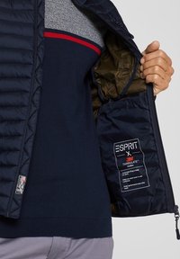 Marineblå quiltet puffer vest, uafgjort for at vise det glatte indvendige for. Har brandingsmærke og 3M Thinsulate isolationsmærke.