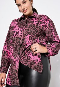 Pink und schwarzes Leopardenmuster-Shirt mit Knopfleiste, langen Ärmeln und lockerer Passform. Schwarze Leggings sind am unteren Ende sichtbar.