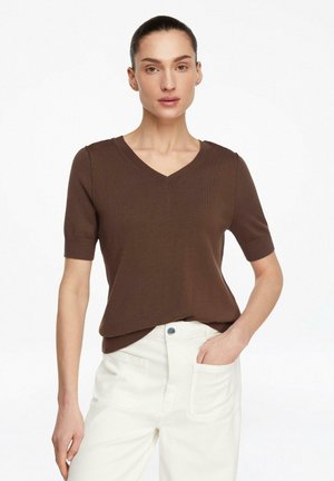 Kobieta w ciemnobrązowej, krótkiej bluzce z dekoltem w serek, wsuniętej w jasne, wysokowcięte spodnie, stojąca z jedną ręką w kieszeni na białym tle.