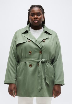 Nő világoszöld öves ballonkabátban, barna gombokkal és arany karika fülbevalóval, egyszínű fehér háttér előtt áll.