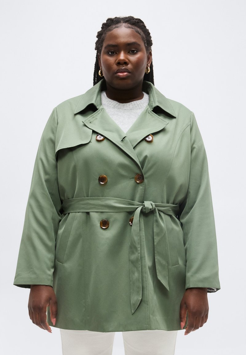 Γυναίκα που φοράει ένα ανοιχτό πράσινο παλτό τύπου trench με ζώνη, καφέ κουμπιά και χρυσούς κρίκους για σκουλαρίκια, στέκεται μπροστά σε ένα απλό λευκό φόντο.