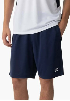 Persona che indossa pantaloncini sportivi blu navy con un logo bianco sulla gamba sinistra e una maglietta bianca, con una mano in tasca.