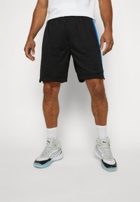 Svarta träningsshorts med blå sidsnederdelar, elastisk midja och lös passform. Kombineras med vita strumpor och ljusgrå sneakers.