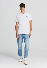 Biała koszulka z krótkim rękawem z nadrukowanym tekstem, w połączeniu z jasnoniebieskimi dżinsami prostymi i białymi trampkami z zielonymi akcentami.