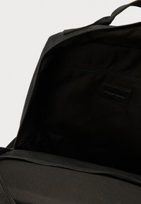 Intérieur de sac à dos en tissu noir avec une poche zippée, texture lisse et une petite étiquette. Design minimaliste avec une mise en page épurée.