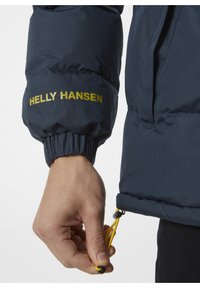 Granatowa kurtka puchowa z elastycznymi mankietami, z żółtymi regulowanymi sznurkami i logo "HELly HANSEN" w złotym kolorze na rękawie.