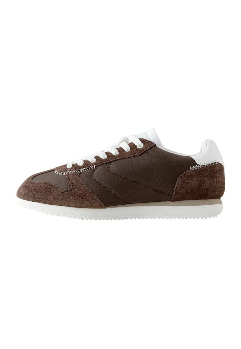 Zapatilla marrón con una combinación de ante y tejido, acentos blancos, suela texturizada y seis ojales para los cordones. Diseño clásico retro.
