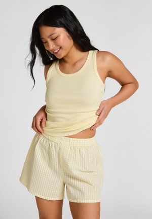 Junge Frau, die ein blassgelbes, geripptes Tanktop justiert, trägt gelb-weiß karierte Shorts, lächelt vor einem schlichten, hellen Hintergrund.