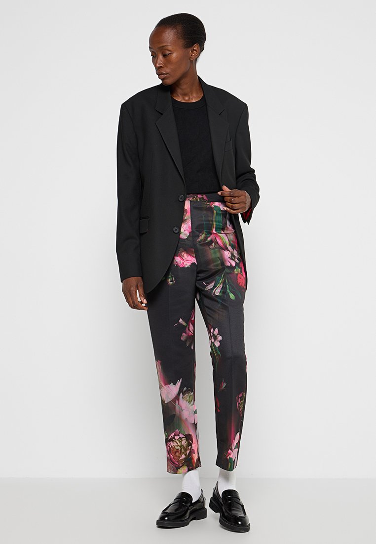 Blazer nero su un top nero, abbinato a pantaloni a fantasia floreale con fiori rosa e verdi su uno sfondo scuro. Scarpe nere lucide.