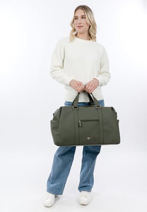 Frau in weißem Pullover und Jeans hält eine große olivgrüne Reisetasche mit vorderer Reißverschlusstasche.